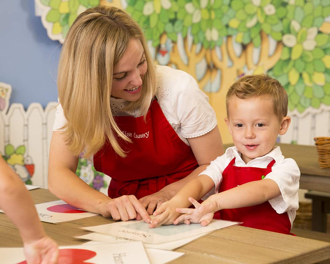 Kids Village teacher in classroom smiling and pointing at shapes on table with preschool students