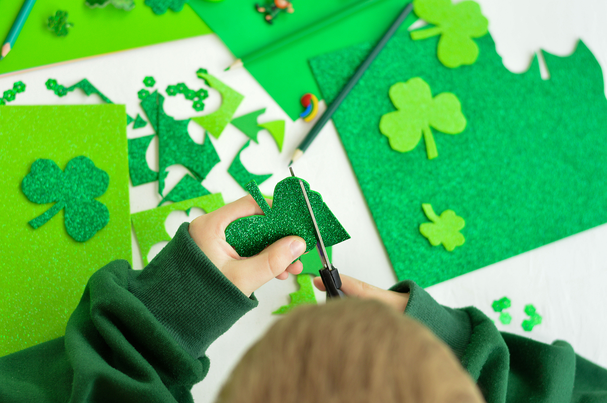 DIY St.Patricks Day decor. A boy cuts with scissors craft card of shiny green paper. Selective focus