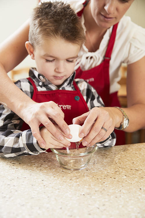 Preschool boy at Kids Village in Orem cracking an egg into a small bowl with a teacher's help