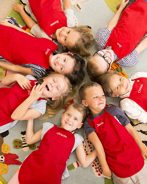 Group of cute private preschool students laying on the ground in a circle with their heads together in the middle while they smile and laugh at the camera